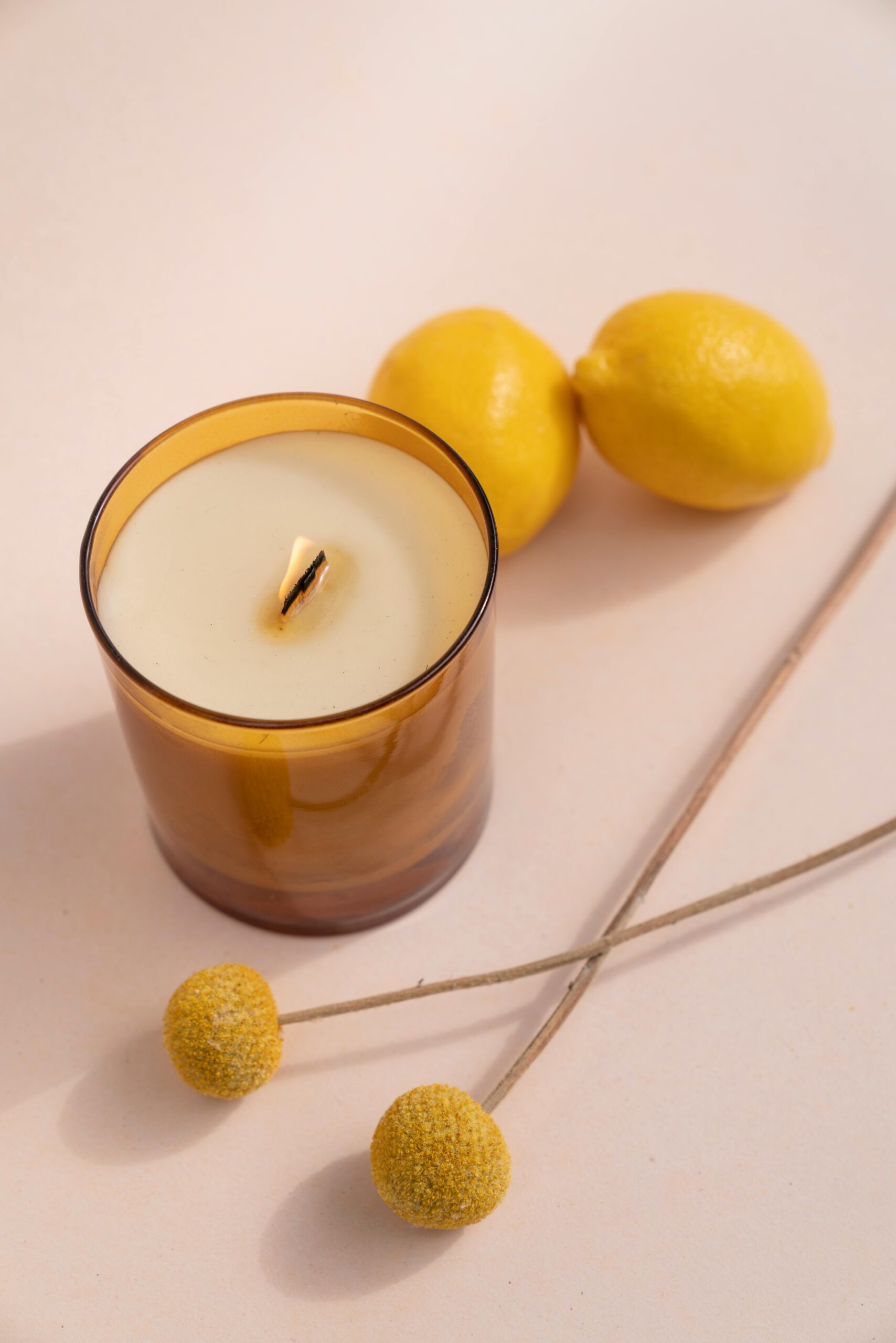 A warm-toned candle beside fresh lemons and craspedia flowers in a minimalist composition.